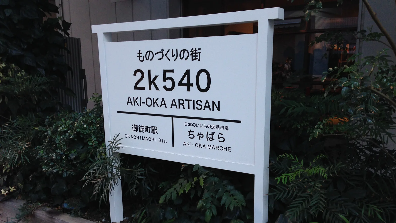 A white 2k540 AKI-OKA ARTISAN sign among plants, photographed in November 2013.